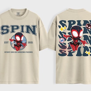 May include: Beige t-shirt featuring a graphic of a superhero character. The front of the shirt has the word "SPIN" above a circular design with the character and the text "EST 2021 Spidey and his amazing friends". The back of the shirt has the word "SPIN" repeated with the character.