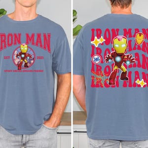 May include: A blue t-shirt featuring the Iron Man character. The front of the shirt has the words "Iron Man" in red, with a graphic of Iron Man in the center. The back of the shirt repeats "Iron Man" in red, with additional graphics.