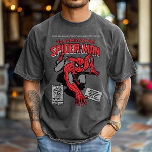 May include: A charcoal grey short-sleeve t-shirt featuring a vintage comic book graphic of Spider-Man in a crouching pose. The text reads: NEVER HAS ANYONE FOUGHT SUCH MERCILESS FOES AS... THE AMAZING SPIDER-MAN, FEATURING PETER PARKER ALIAS SPIDER-MAN.