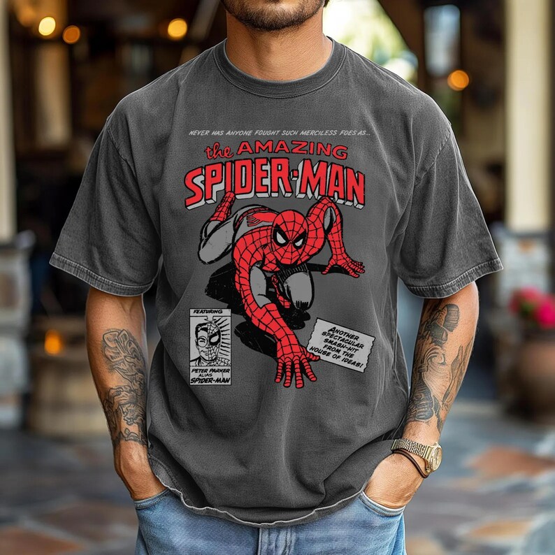 May include: A charcoal grey short-sleeve t-shirt featuring a vintage comic book graphic of Spider-Man in a crouching pose. The text reads: NEVER HAS ANYONE FOUGHT SUCH MERCILESS FOES AS... THE AMAZING SPIDER-MAN, FEATURING PETER PARKER ALIAS SPIDER-MAN.