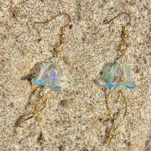 May include: A pair of jellyfish earrings with gold-colored hooks and tendrils. The jellyfish bodies are translucent with a blue rim, and the tendrils are adorned with small, clear beads. The earrings are set against a sandy background.