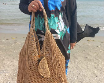 Könnte beinhalten: Eine große, offen gewebte, hellbraune Häkeltasche mit langen Fransen und einem blattförmigen Akzent. Die Tasche hat stabile Griffe und wird von einer Person an einem Sandstrand gehalten. Die Tasche ist ein stilvolles Accessoire.