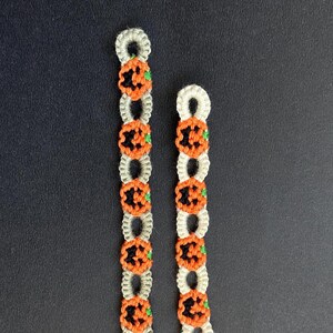 Puede incluir: Dos pulseras hechas a mano con temática de Halloween. Cada pulsera presenta un patrón repetido de formas de calabaza naranja con detalles negros y tallos verdes, conectadas por un cordón gris claro o blanco. Las pulseras se muestran sobre un fondo oscuro.