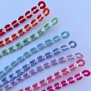 Puede incluir: Una colección de pulseras de ganchillo coloridas. Cada pulsera presenta una cadena de formas ovaladas con pequeñas flores blancas y un lazo en el extremo. Las pulseras son de color rojo, naranja, verde, azul, morado, rosa y fucsia.