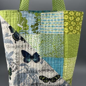Könnte beinhalten: Gesteppte Tragetasche mit Patchwork-Design. Die Tasche hat verschiedene grüne und blaue Blumen- und Schmetterlingsmuster. Die Griffe sind grün mit weißen Punkten. Auf der Tasche stehen die Wörter "BORBOLETA" und "QUERCUS SUBUS".