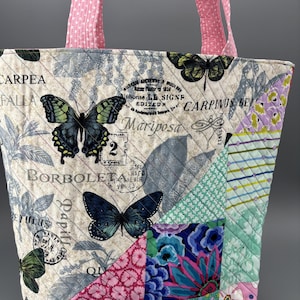 Könnte beinhalten: Gesteppte Tragetasche mit Schmetterlings- und Pflanzenmuster. Die Tasche hat ein Patchwork-Design mit verschiedenen Stoffen in Creme-, Blau-, Rosa- und Grüntönen. Rosa gepunktete Griffe runden das Design ab.
