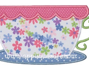 May include: A pink and white teacup with a floral pattern and a green handle. The teacup is sitting on a blue and white saucer with white polka dots. The text "www.appliquecafe.com" is printed on the saucer.