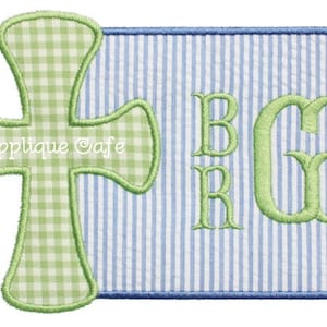 May include: A green cross with a gingham pattern is stitched onto a blue and white striped fabric background. The fabric has the letters "BRG" stitched in green on it. The text "Applique Cafe" is stitched in white on the cross.