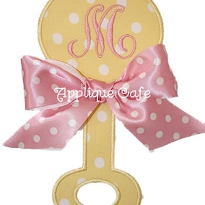 Peut inclure: Applique en forme de hochet jaune à pois blancs et nœud en satin rose. Le hochet présente une lettre "M" rose brodée et le texte "Applique Cafe".