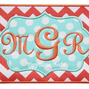 Op de afbeelding: Een turquoise stof met witte stippen en een gegolfde rand, met het monogram "M G R" in oranje borduurwerk. De stof is omlijst door een rood en wit zigzagpatroon. De tekst "Applique Cafe" is in groen onder het monogram geborduurd.