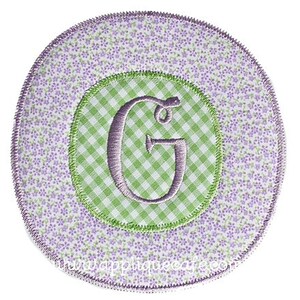 Könnte beinhalten: Eine runde Applikation aus lila und grünem Stoff mit einem weißen Buchstaben G in der Mitte. Die Applikation hat ein Karomuster und ein Blumenmuster.