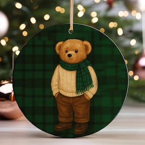 May include: Round Christmas ornament featuring a teddy bear wearing a cream-colored sweater, green scarf, and brown pants. The bear stands against a green plaid background. The ornament hangs from a gold string, with Christmas tree decorations in the background.