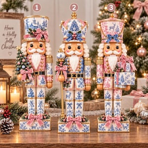 Op de afbeelding: Drie decoratieve notenkraker-beeldjes in een roze en blauw kleurenpalet met patronen van sneeuwvlokken, herten en bomen. Elke notenkraker staat op een rechthoekige voet en is versierd met strikken, gouden accenten en wit kunsthaar.