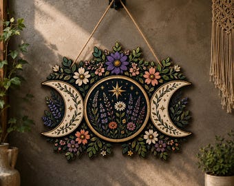 May include: A decorative wall hanging featuring a central circular panel with a star and wildflowers, flanked by two crescent moons. The piece is adorned with colorful floral motifs in shades of purple, pink, and white, all suspended by a rope.