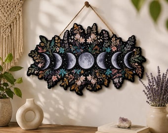 May include: A horizontal wall hanging featuring a series of seven moon phases arranged in a row, surrounded by a dense arrangement of colorful flowers and foliage in shades of pink, blue, and green, suspended by a thin brown twine cord.