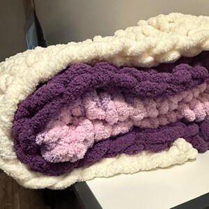 May include: A folded chunky knit blanket in cream, purple, and pink. The layers of the blanket are visible, highlighting the different colours and textures. The thick yarn gives a cosy and inviting appearance.