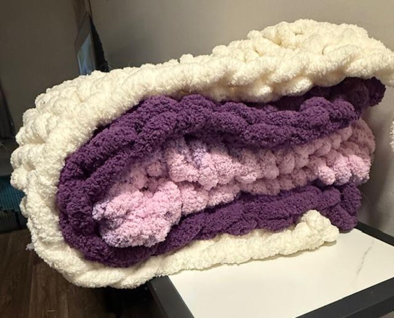May include: A folded chunky knit blanket in cream, purple, and pink. The layers of the blanket are visible, highlighting the different colours and textures. The thick yarn gives a cosy and inviting appearance.