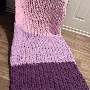 May include: A colour block chunky knit blanket. The blanket is made with thick yarn and features sections of white, light pink, lavender, and deep purple. The textured blanket is designed for comfort and home decor.