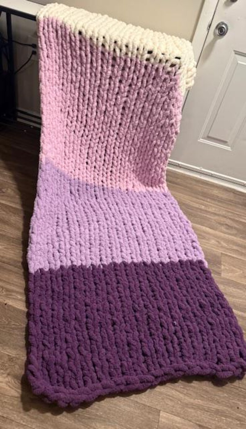May include: A colour block chunky knit blanket. The blanket is made with thick yarn and features sections of white, light pink, lavender, and deep purple. The textured blanket is designed for comfort and home decor.