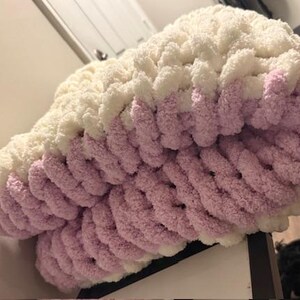 May include: A chunky knit blanket in cream and lavender. The blanket is made of thick, soft yarn and features a two-tone design. The top portion is cream, transitioning to lavender at the bottom. The blanket is folded, showing the texture of the knit.
