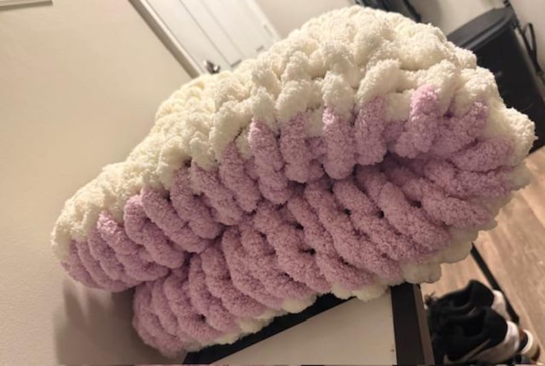 May include: A cream and lavender chunky knit blanket. The blanket is made from thick, soft yarn and has a two-tone design. The top section is cream, fading into lavender at the bottom. The blanket is folded, displaying the texture of the knit, which is approximately 100 cm wide.