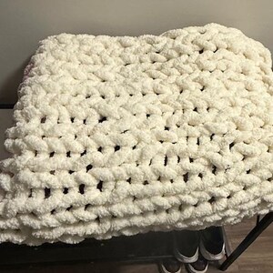 May include: A cream-coloured, chunky knit blanket. The blanket is made of thick, soft yarn, with a visible woven texture. The blanket is folded and appears to be resting on a dark surface. The overall look is cosy and inviting.