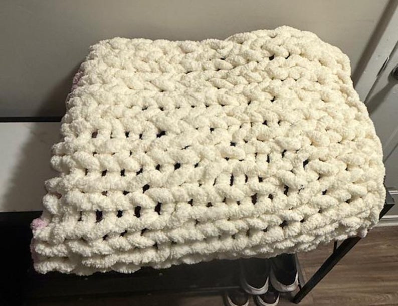 May include: A cream-coloured, chunky knit blanket. The blanket is made of thick, soft yarn, with a visible woven texture. The blanket is folded and appears to be resting on a dark surface. The overall look is cosy and inviting.