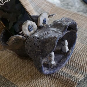 May include: A ceramic hippo-shaped phone holder in shades of blue and gray. The hippo has large, expressive eyes with blue irises and a wide open mouth. The phone rests in the hippo's mouth. The holder is textured with small holes.