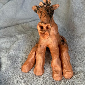 Op de afbeelding: Een handgemaakt terracotta beeld van een giraffe met een getextureerd oppervlak en zwarte markeringen. De giraffe zit met zijn poten uitgestrekt en zijn hoofd omhoog gekanteld.