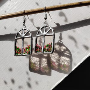 Puede incluir: Un par de pendientes plateados con forma de ventana. Cada pendiente presenta un cristal transparente con un diseño floral de flores rosas y verdes en la parte inferior. Los pendientes están suspendidos de un palillo de madera, proyectando sombras sobre una superficie blanca.