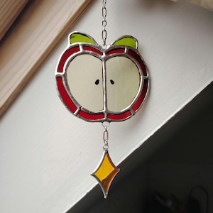 Peut inclure: Un attrape-soleil en vitrail en forme de pomme coupée avec une bordure rouge, un centre jaune pâle et deux petites graines noires. Deux petites feuilles vertes sont situées au sommet. Un losange en verre ambré pend en bas sur une chaîne.
