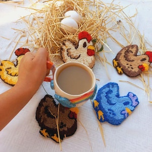 Pode incluir: Uma caneca de cerâmica com um design de arco-íris e uma chávena de café, rodeada por cinco bases bordadas em forma de galinha. Um ninho de palha com dois ovos brancos está no fundo. As bases são amarelas, castanhas e azuis.