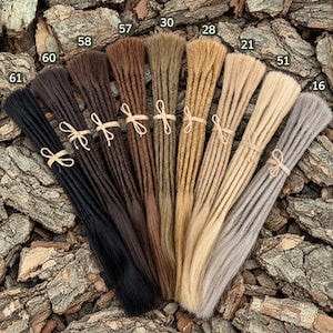 Peut inclure: Une collection de dix paquets de dreadlocks en cheveux synthétiques disposés en éventail, allant du noir au brun foncé, brun clair, blond et gris. Chaque paquet est attaché avec une ficelle beige clair et numéroté de 16 à 61.
