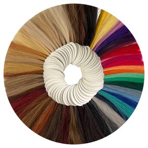 Puede incluir: Un abanico circular de muestras de color de cabello que muestra una variedad de colores de cabello. Los colores van desde el rubio y el marrón hasta tonos vibrantes como el rojo, el morado y el verde azulado. El abanico está sujeto por un soporte circular blanco.