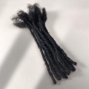 Può includere: Un fascio di extension per capelli neri acconciate come dreadlock individuali. Le ciocche hanno un aspetto testurizzato e opaco, con un colore scuro uniforme su tutta la superficie. Le ciocche sono raggruppate in alto e si aprono leggermente verso il basso.