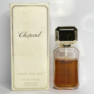 May include: A rectangular off-white perfume box with gold trim and the text Chopard Happy Diamonds Eau de Parfum Vaporisateur Natural Spray, next to a clear glass perfume bottle with a gold faceted cap containing amber liquid.
