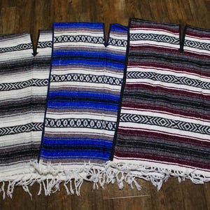 May include: Three colorful serape ponchos with fringe. The ponchos are in shades of gray, blue, and burgundy, with horizontal stripes and geometric patterns. They are displayed on a wooden surface.