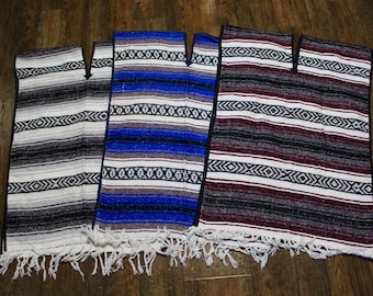 May include: Three colorful serape ponchos with fringe. The ponchos are in shades of gray, blue, and burgundy, with horizontal stripes and geometric patterns. They are displayed on a wooden surface.
