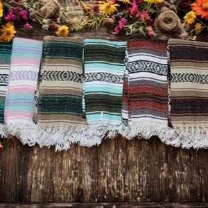 May include: Six folded woven blankets featuring traditional striped patterns in various color combinations including teal, pink, olive green, light blue, rust red, and brown, all finished with white knotted fringe along the bottom edges.