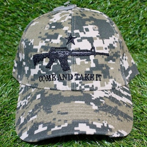 Op de afbeelding: Camouflage baseball cap met een zwarte afbeelding van een geweer en een ster, met de tekst "COME AND TAKE IT" eronder geborduurd. De cap heeft een gebogen klep en een stoffen band. Het patroon is in groen, bruin en beige.