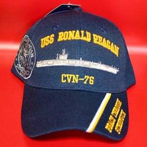 Könnte beinhalten: Marineblaue Baseballkappe mit gestickten Details. Sie zeigt "USS RONALD REAGAN" in Gelb, eine Schiffillustration und "CVN-76". Auf der Seite befindet sich ein Aufnäher und Text auf dem Schirm.