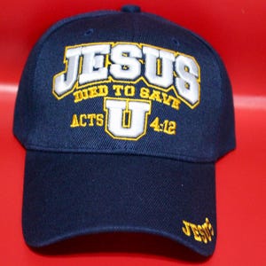 May include: Navy blue baseball cap with embroidered text. The text includes "JESUS" in white with a yellow outline, "DIED TO SAVE", "ACTS U 4:12", and "Jesus" on the side. The cap has a curved brim and a rounded crown.