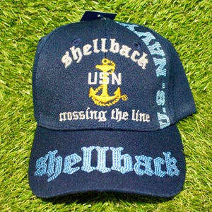 Peut inclure: Casquette de baseball bleu marine avec texte brodé et un emblème d'ancre doré. La casquette porte les mots "Shellback", "USN" et "crossing the line". Le bord porte également le texte "U.S. NAVY".