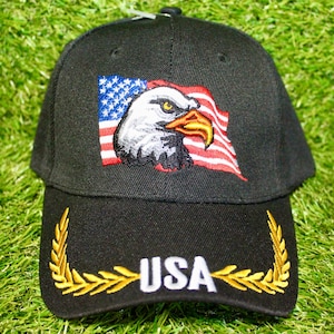 May include: Black baseball cap featuring an embroidered American flag and a bald eagle design. The cap's front displays the word "USA" in white, framed by golden laurel leaves. A patriotic accessory.