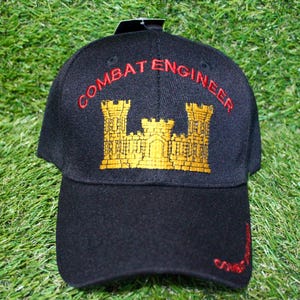 May include: Black baseball cap with "COMBAT ENGINEER" embroidered in red, and a gold emblem of a castle. The cap has a curved brim and is set against a green background.