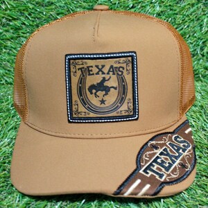May include: A brown trucker cap with a mesh back and a curved peak. The front displays a square patch with the word "TEXAS" and a cowboy on a horse. The peak also has the word "TEXAS" embroidered on it.