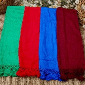 Op de afbeelding: Vier kleurrijke sjaals in groen, rood, blauw en bordeaux worden getoond. Elke sjaal heeft een decoratieve franje langs de onderkant. De sjaals zijn naast elkaar geplaatst op een getextureerd, neutraal oppervlak, met manden en bloemen op de achtergrond.