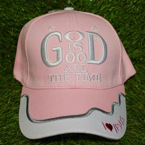 Könnte beinhalten: Rosa Baseballkappe mit dem gestickten Text "GOD IS GOOD ALL THE TIME" in Silber. Der Schirm hat einen weißen Rand mit dem Text "I ❤️ Jesus" und einem roten Herz.