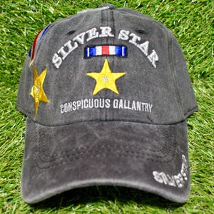May include: A charcoal gray baseball cap with the words "SILVER STAR" embroidered in white above a yellow star. The text "CONSPICUOUS GALLANTRY" is embroidered below the star. A small ribbon and a gold star are on the side.