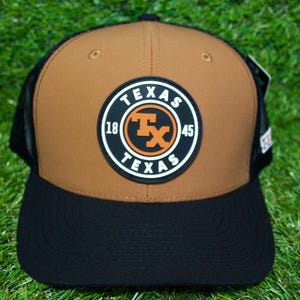 Może przedstawiać: Brązowo-czarna czapka typu trucker z okrągłą naszywką z napisem "TEXAS" i pomarańczowymi literami "TX". Czapka ma siatkowy tył i zakrzywiony daszek.